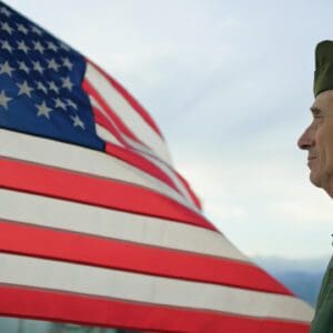 Veteran in uniform beside waving American flag.